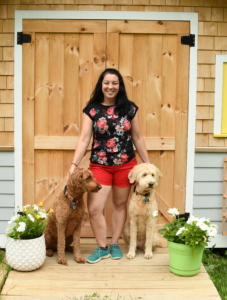 dark haired woman standing in front of doorway with two goldendoodles next to her