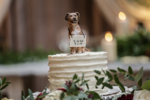 clay figurine of a tan dog sitting on top of a wedding cake with "I do too" written on a sign hanging from it's neck